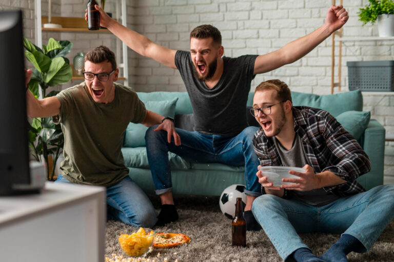 EnParlons.com - Trois amis supporters enthousiastes célébrant avec énergie devant leur télévision dans un salon moderne : un homme en t-shirt vert olive à gauche levant les bras, un homme au centre en t-shirt gris foncé debout sur le canapé turquoise brandissant une bouteille et criant de joie, et un homme à droite en chemise à carreaux tenant un bol de pop-corn, avec ballon de football, bières et snacks (chips, pizza) au sol, illustrant l'ambiance conviviale du visionnage de matchs sportifs via les meilleurs sites IPTV 2025 en France